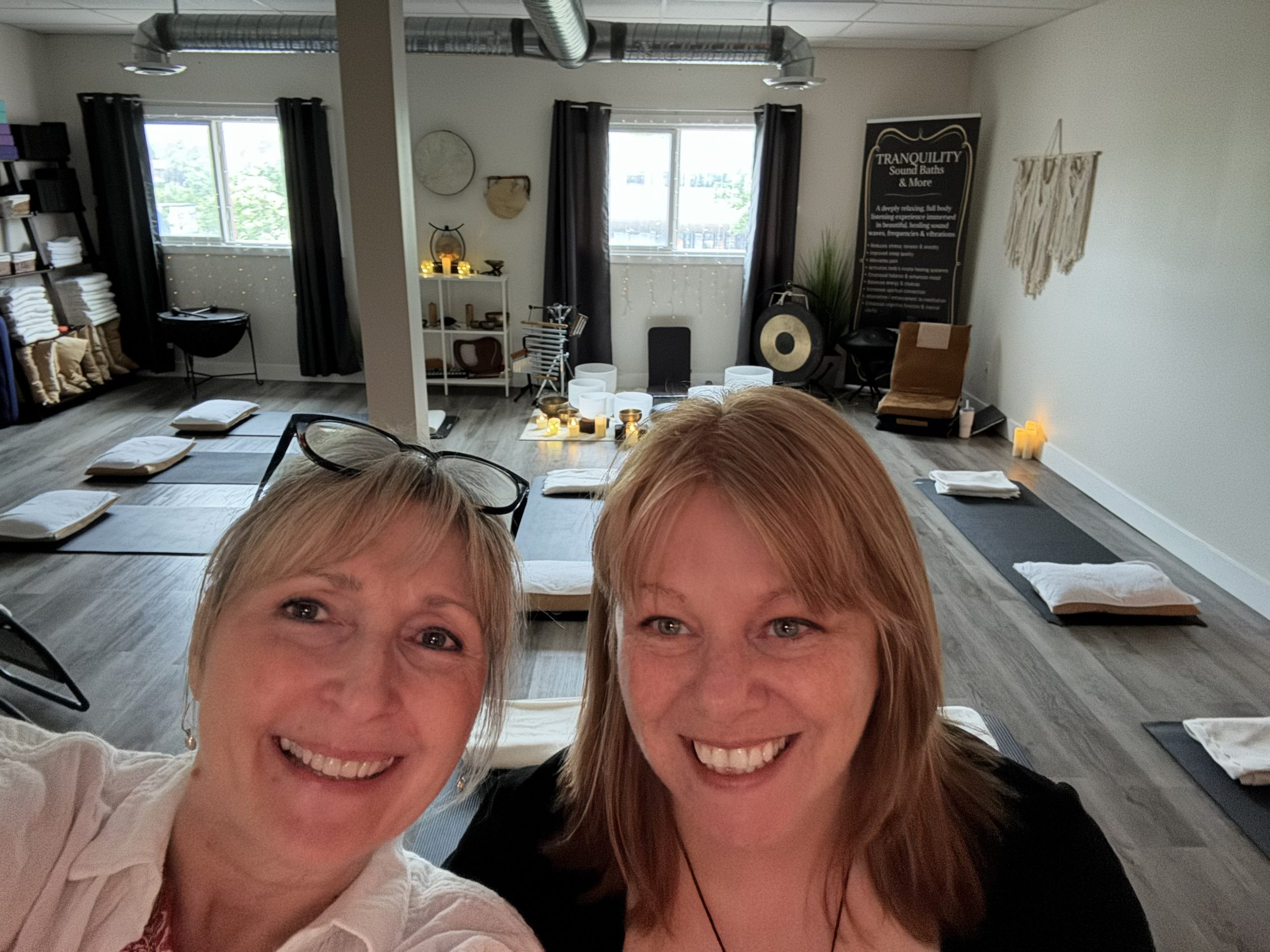 Two facilitators smile in front of a sound bath setup with bowls, candles, and floor mats.